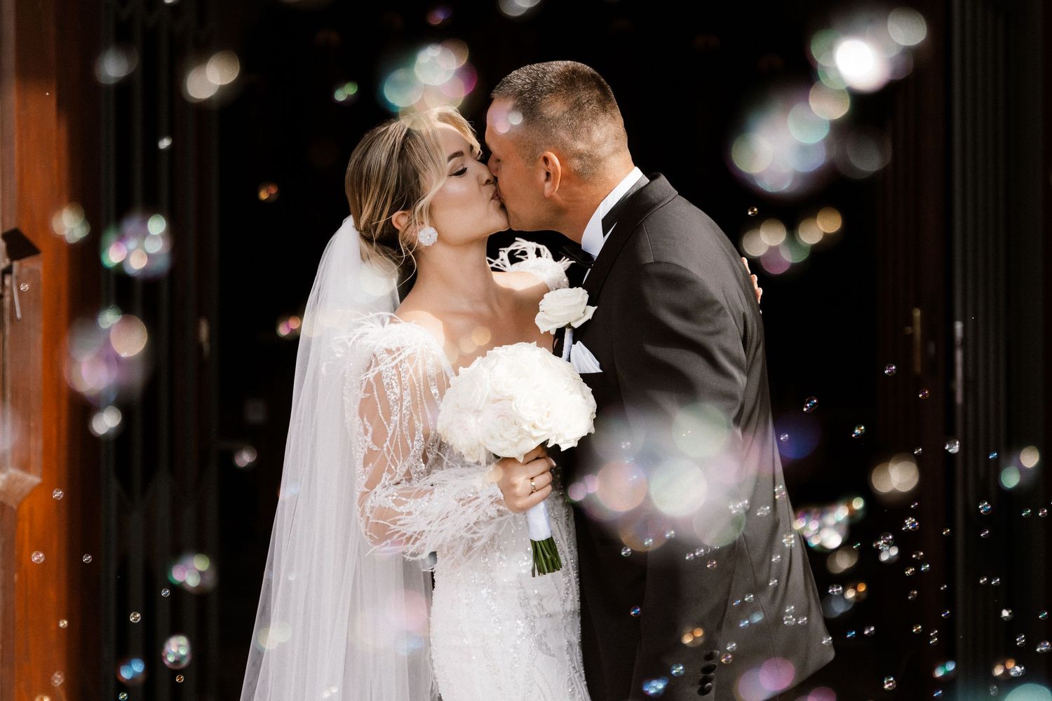 Bride and groom share a romantic kiss surrounded by floating bubbles, bride holding white floral bouquet in wedding attire.