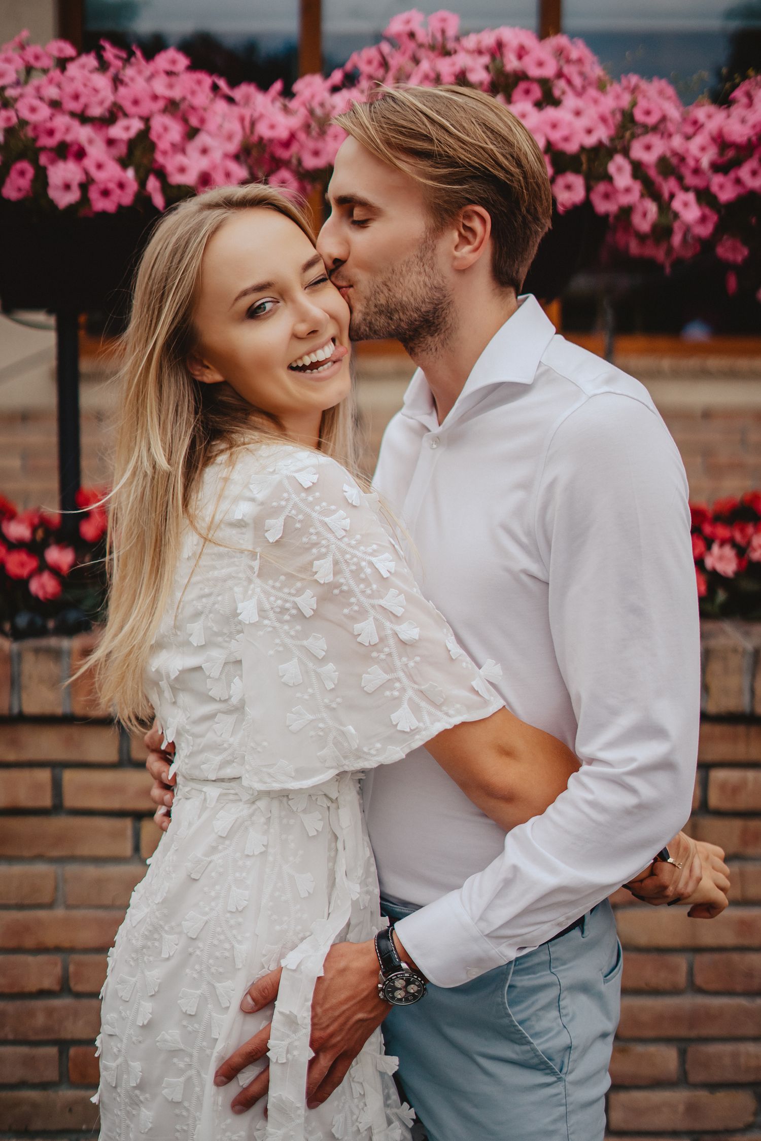 Romantic couple embracing against brick wall with pink flowers blooming in background.