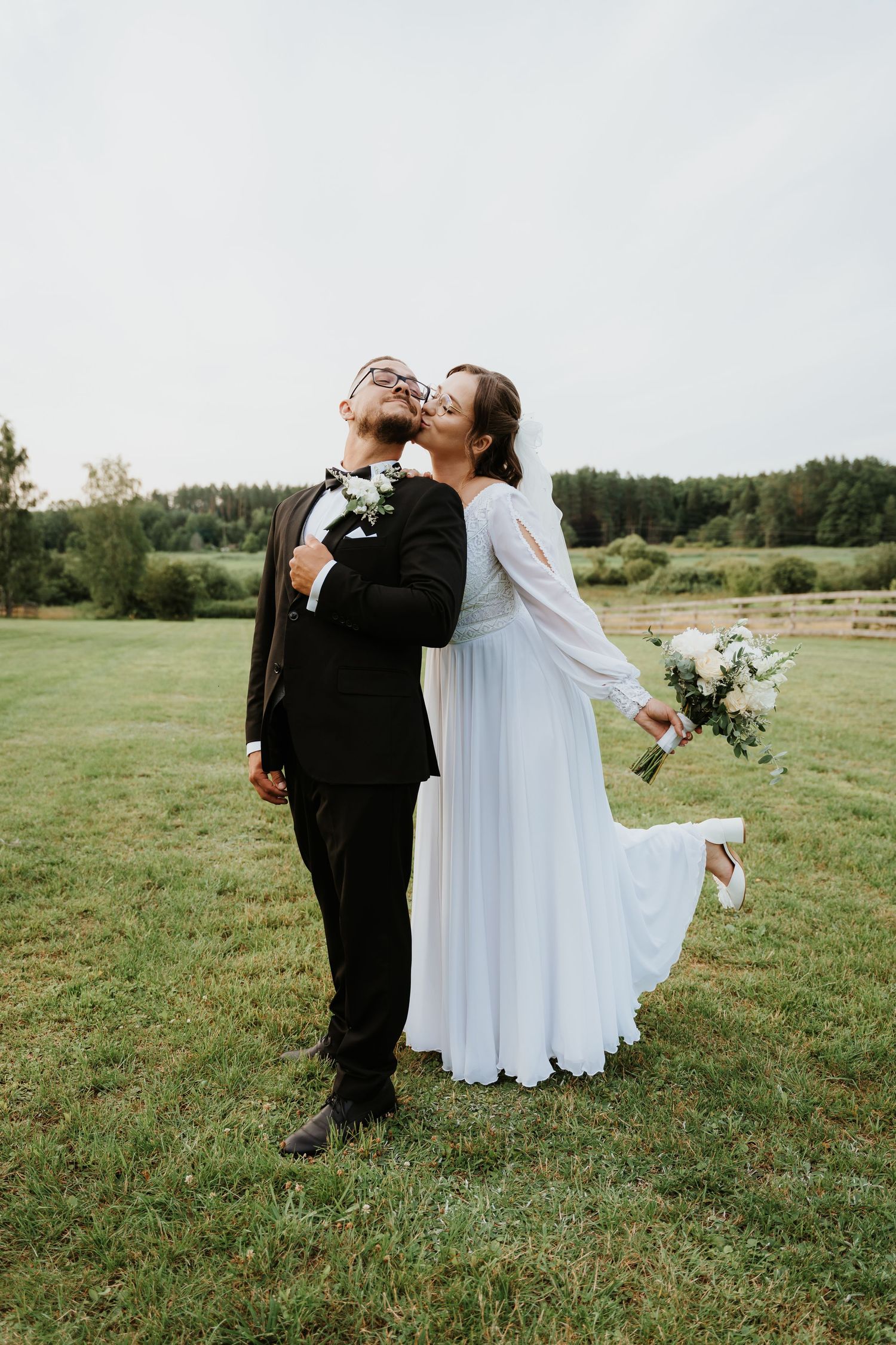 Bride in white gown kisses groom in black suit outdoors on green grass field with bouquet of white flowers.