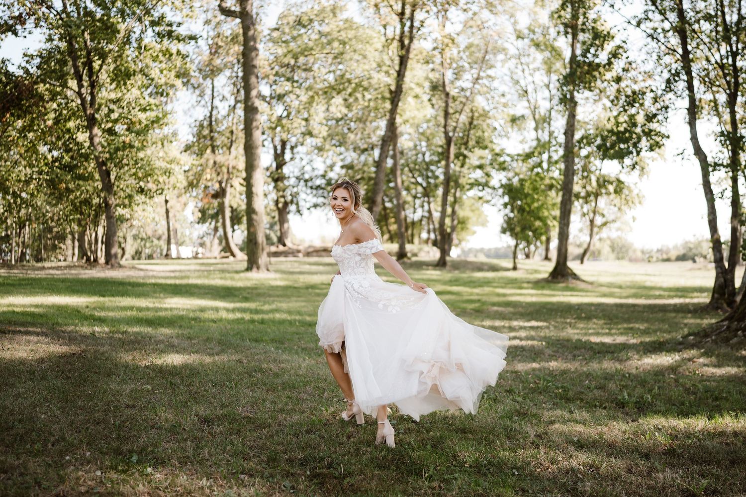 A bride in a flowing white gown poses gracefully in a sunlit wooded area during an outdoor wedding photoshoot.