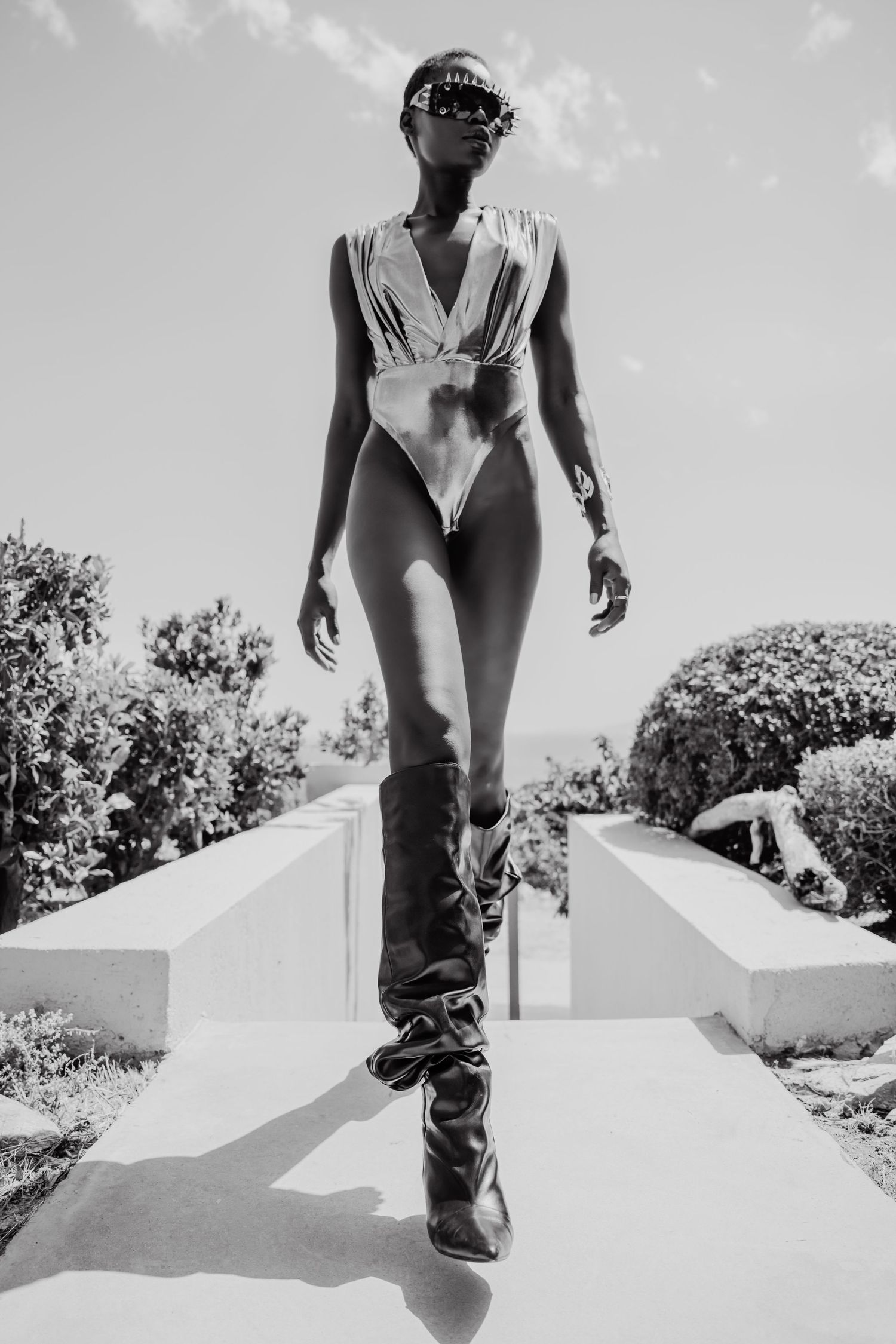 Black and white photo of a woman in a bodysuit and tall boots posing outdoors on a sunny day.