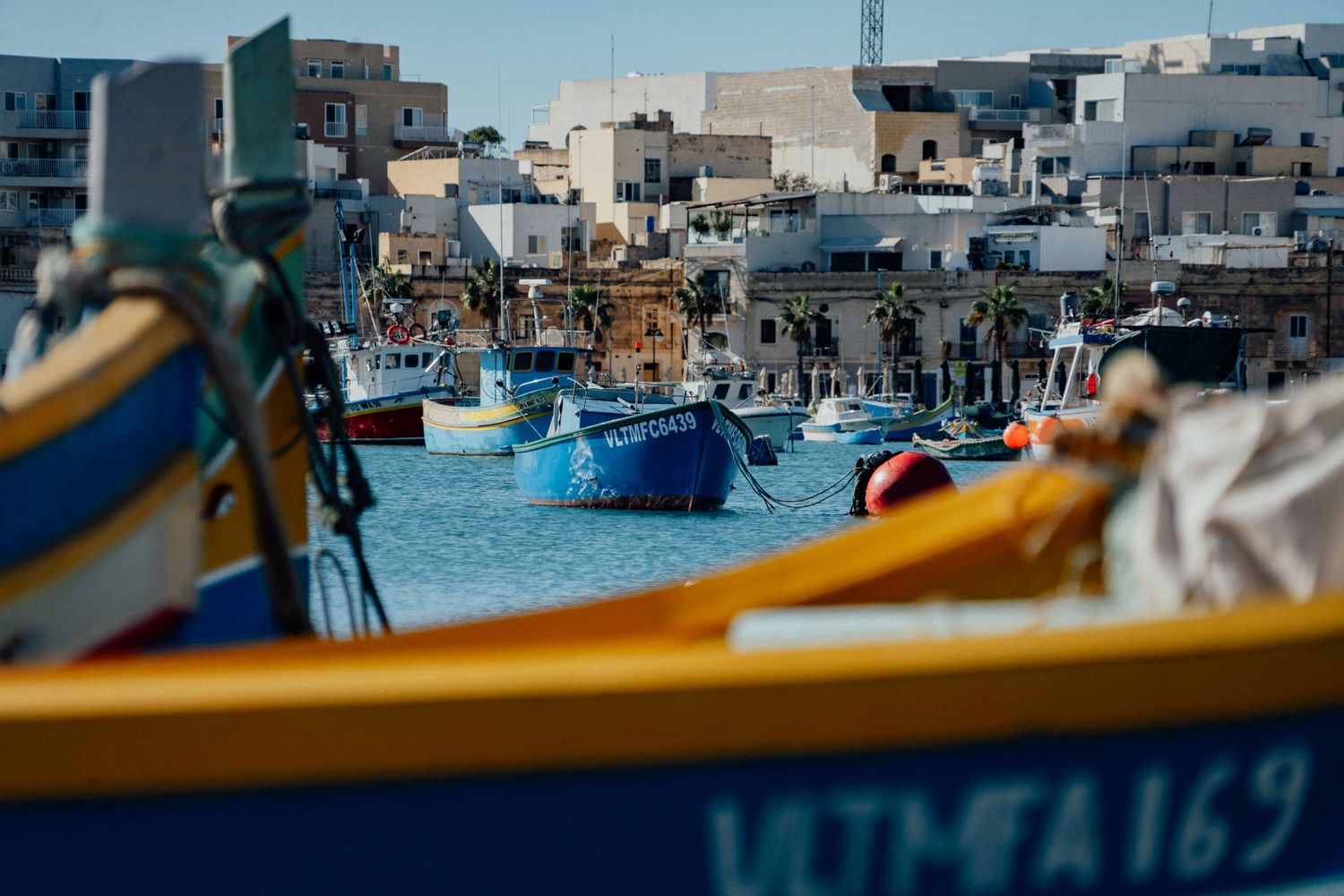 Traditional colorful fishing boats lined up in a Mediterranean harbor with buildings in the background.