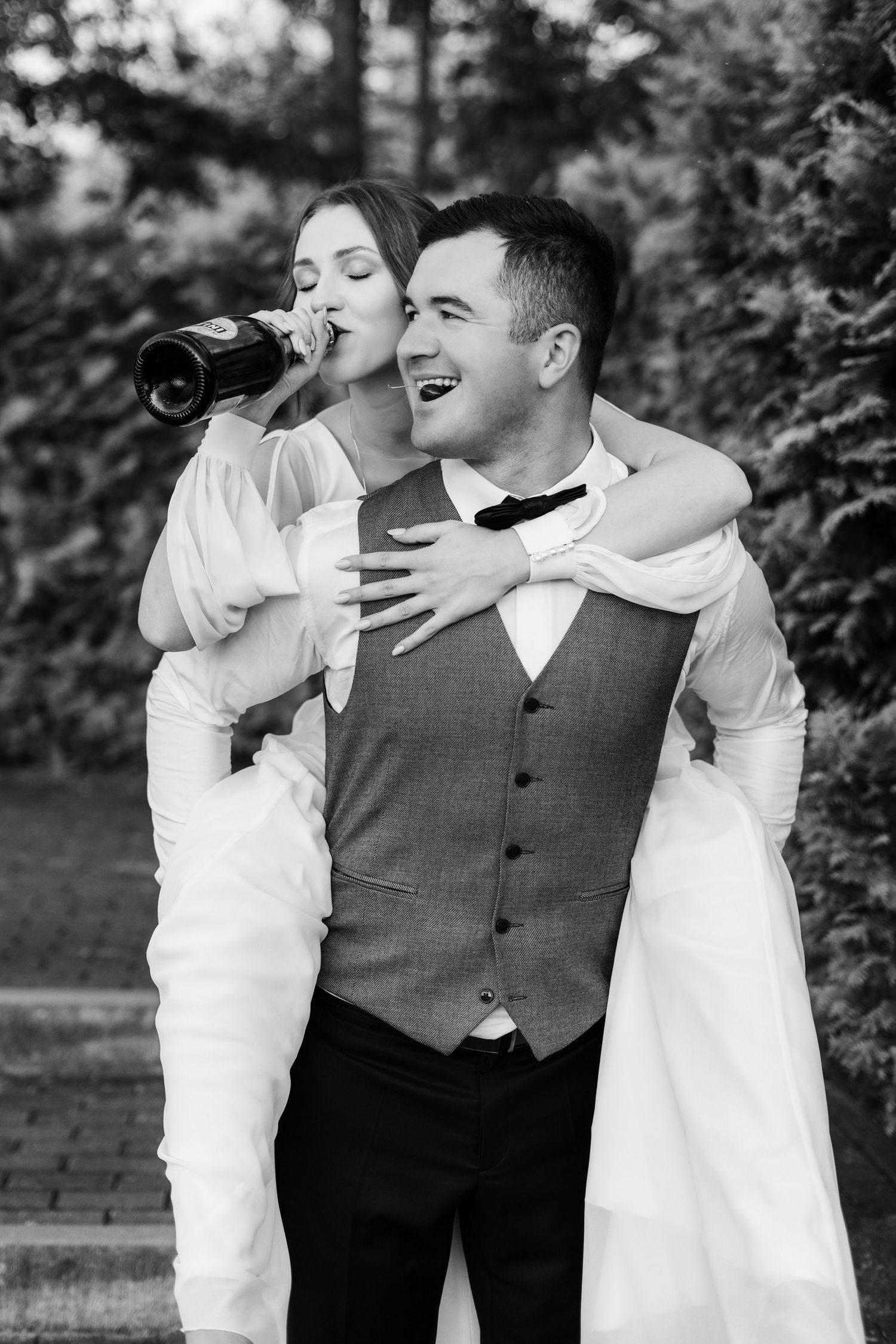 A black and white wedding photo showing a couple sharing a fun moment as the bride drinks from a bottle.