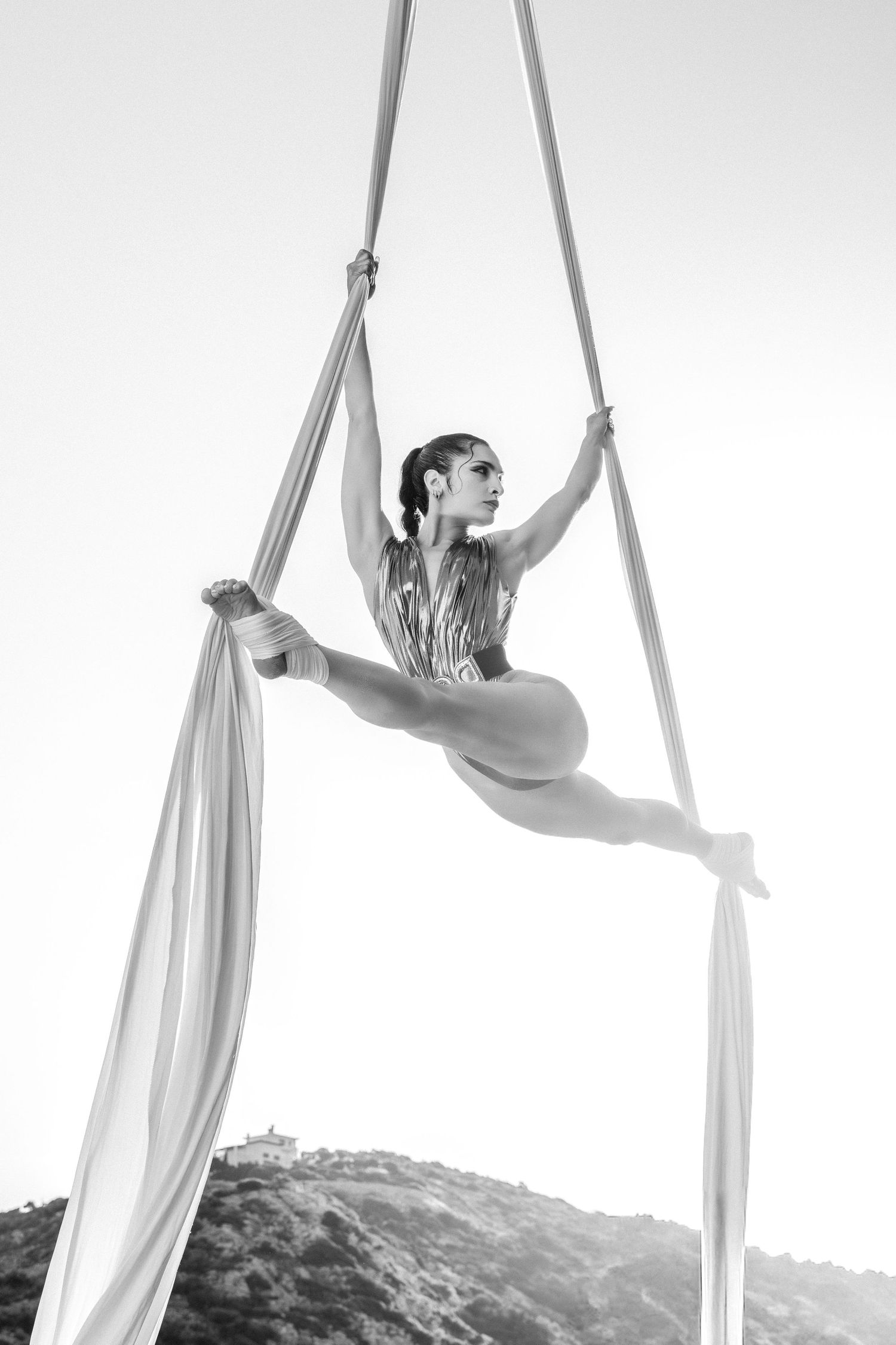 Black and white photo of a female aerialist performing a split on aerial silks outdoors against a bright sky.