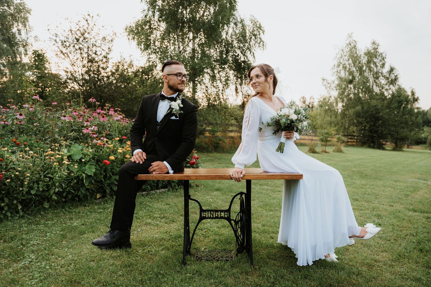 Bride and groom sitting apart on a vintage sewing table in a lush garden, bride holding a bouquet.