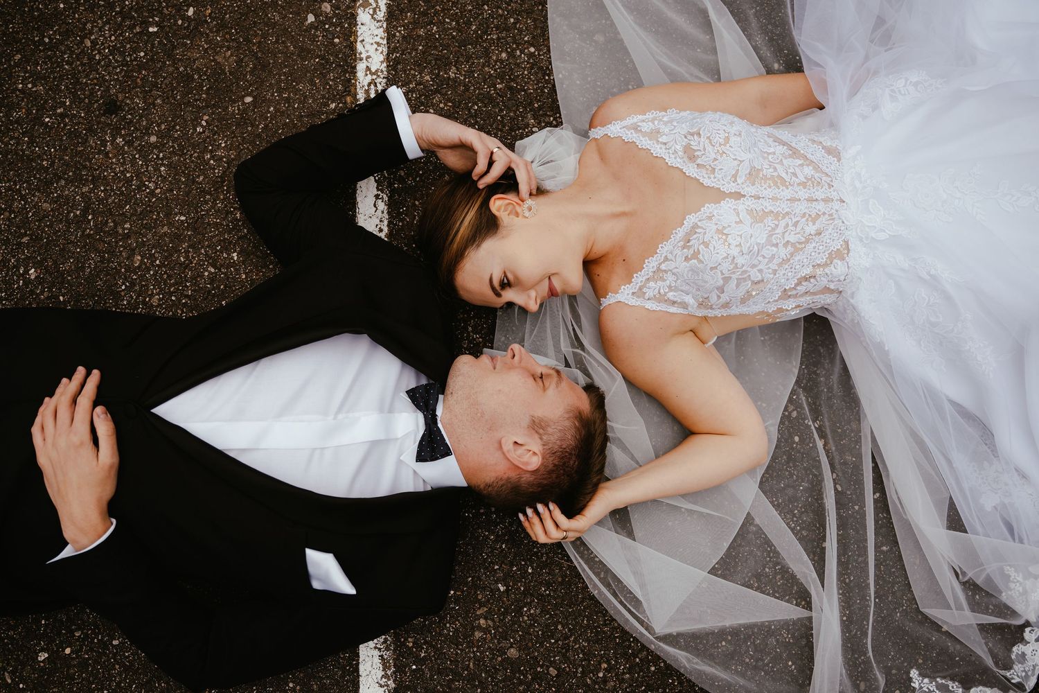 A romantic wedding couple lies on dark ground while sharing an intimate moment in formal attire.