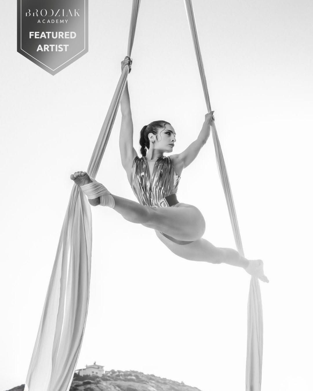 Black and white photo of an aerial silk performer doing a split mid-air, featured artist at Alo Dziak Academy.