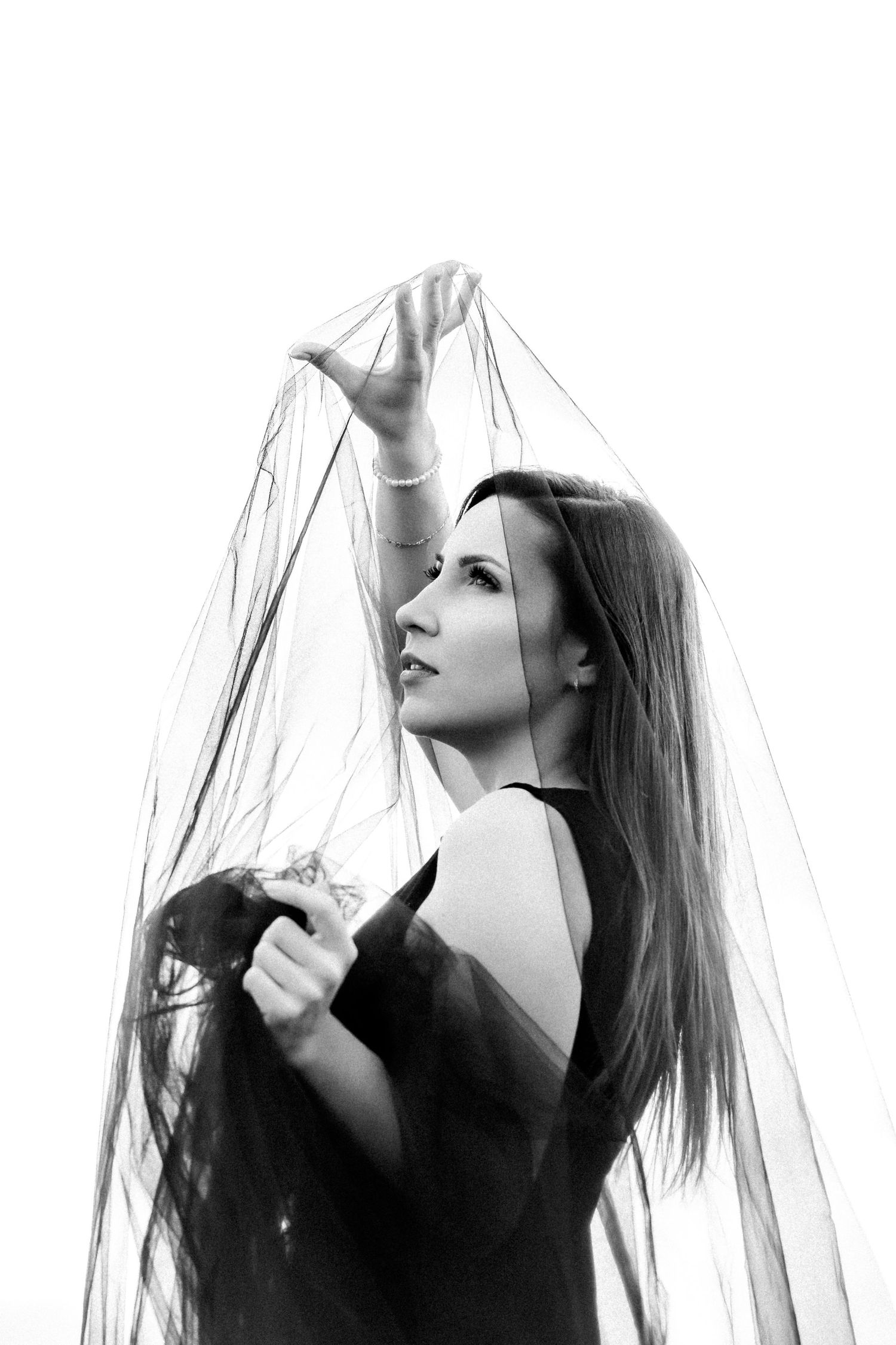 Black and white photo of a woman in dark dress holding sheer fabric veil above her head against white background.