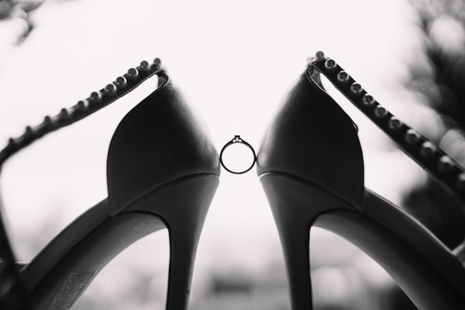 Wedding ring balanced between two black high heel shoes in artistic black and white photography.