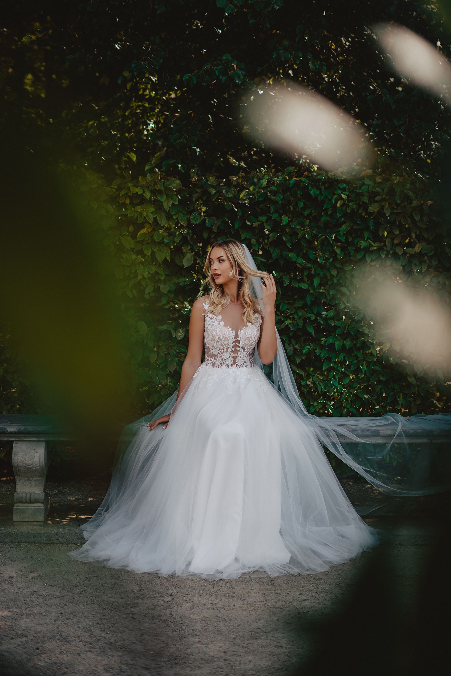 Bride in flowing white wedding dress sitting in garden setting with artistic bokeh effects.