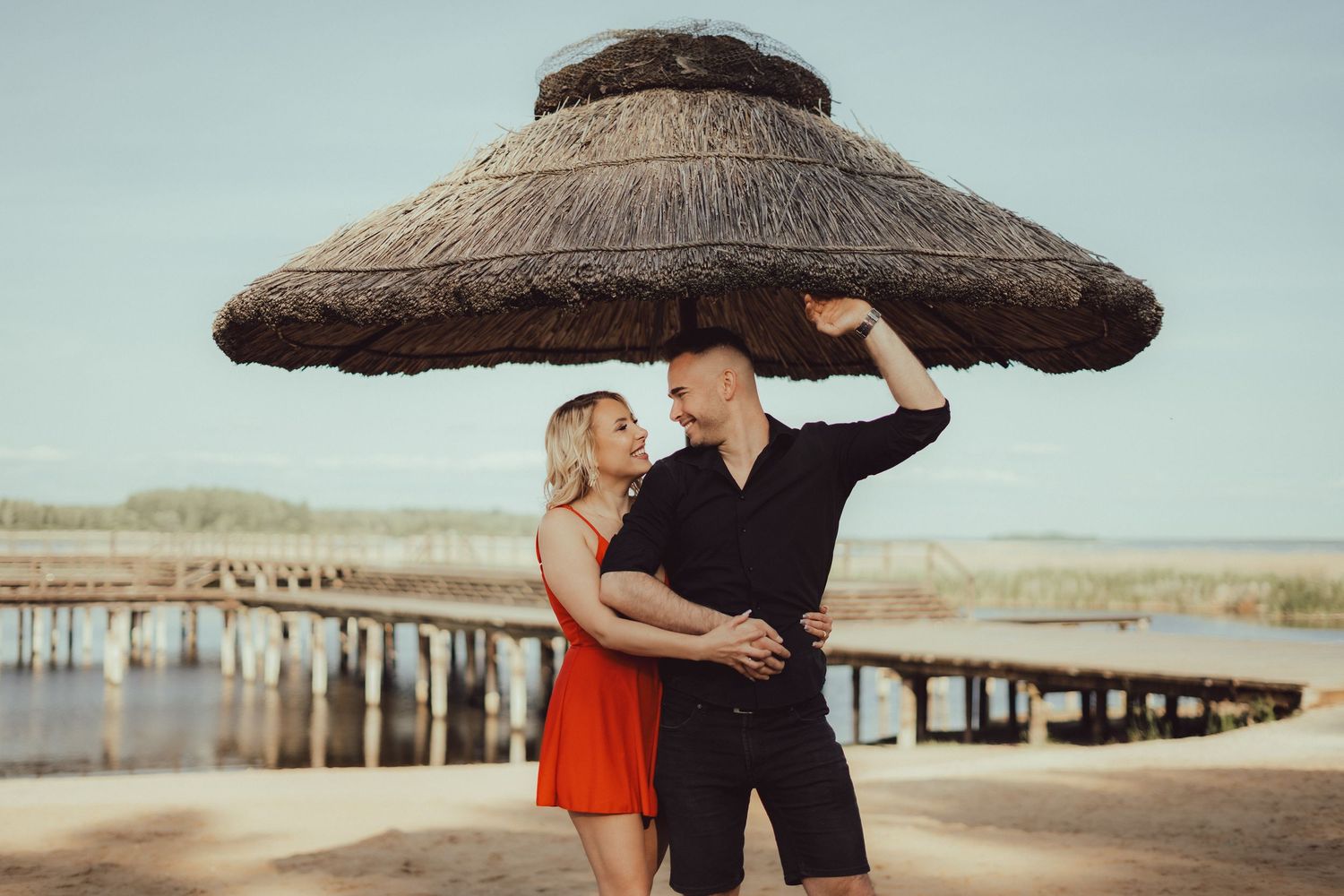A couple embraces on a sandy beach during sunset with a wooden pier in the background.