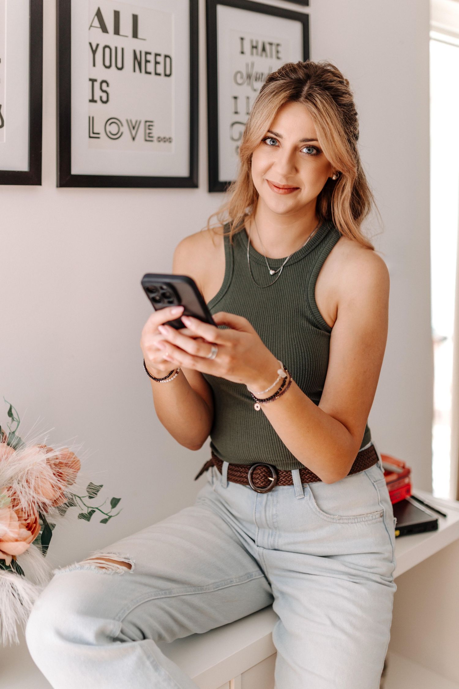 Young blonde woman in olive tank top and jeans holding smartphone, sitting in modern room with motivational wall art.
