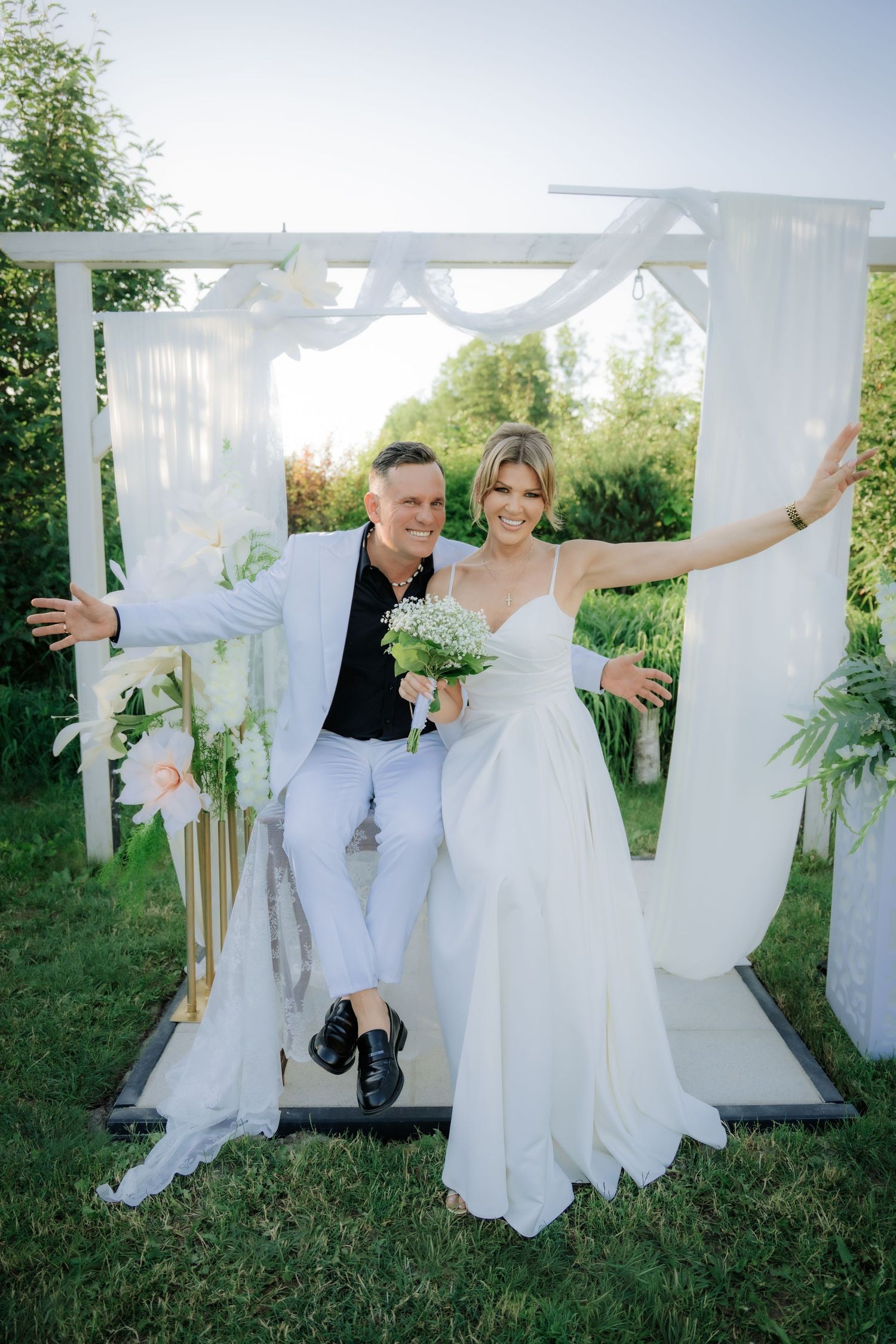 Happy bride and groom posing under white wedding arch outdoors, bride holding bouquet with arms outstretched.