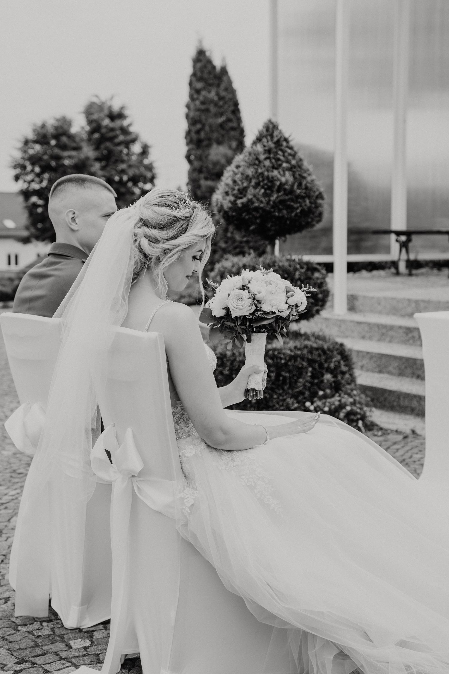 Black and white wedding photo showing a bride in a flowing dress sitting on steps holding a bouquet outdoors.
