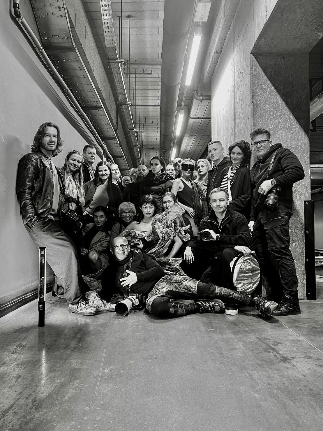Black and white group photo of people posing together in an industrial hallway with fluorescent lighting.