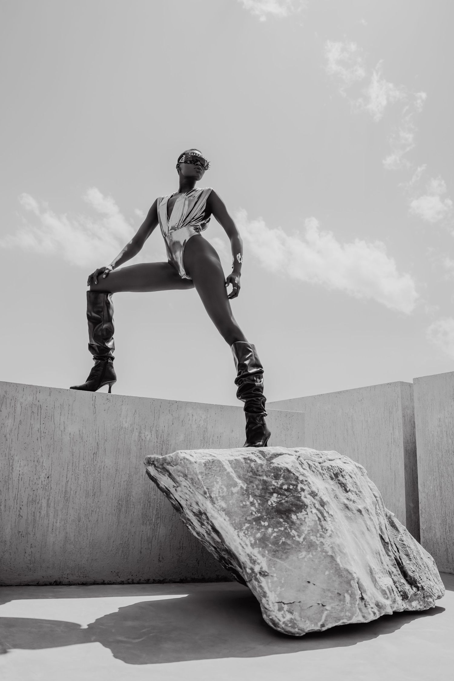 Black and white photo of a model posing dramatically on a rock in a bodysuit and boots under a cloudy sky.