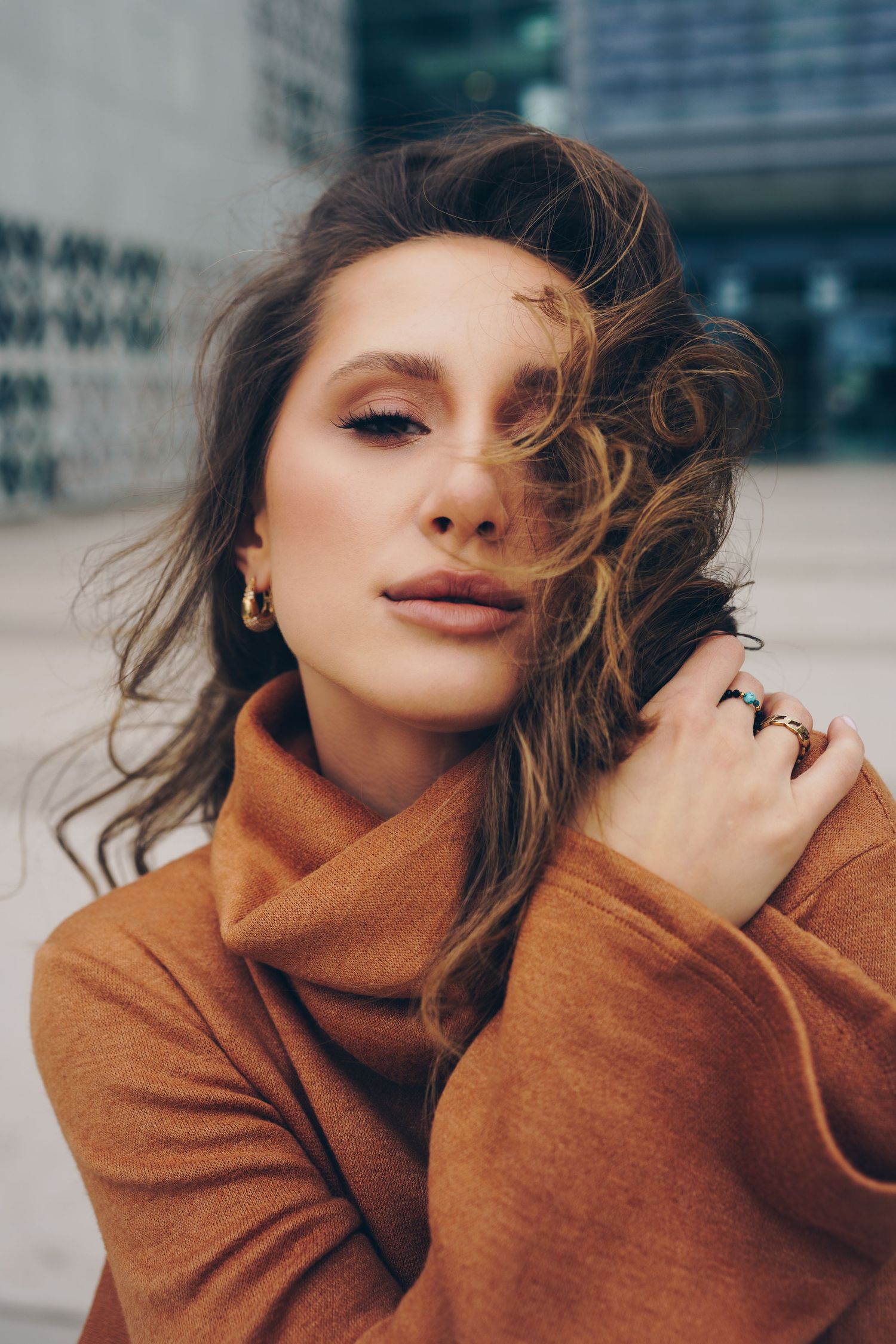 Young woman in brown turtleneck coat with wavy hair blowing, wearing gold jewelry, posing outdoors near modern building.