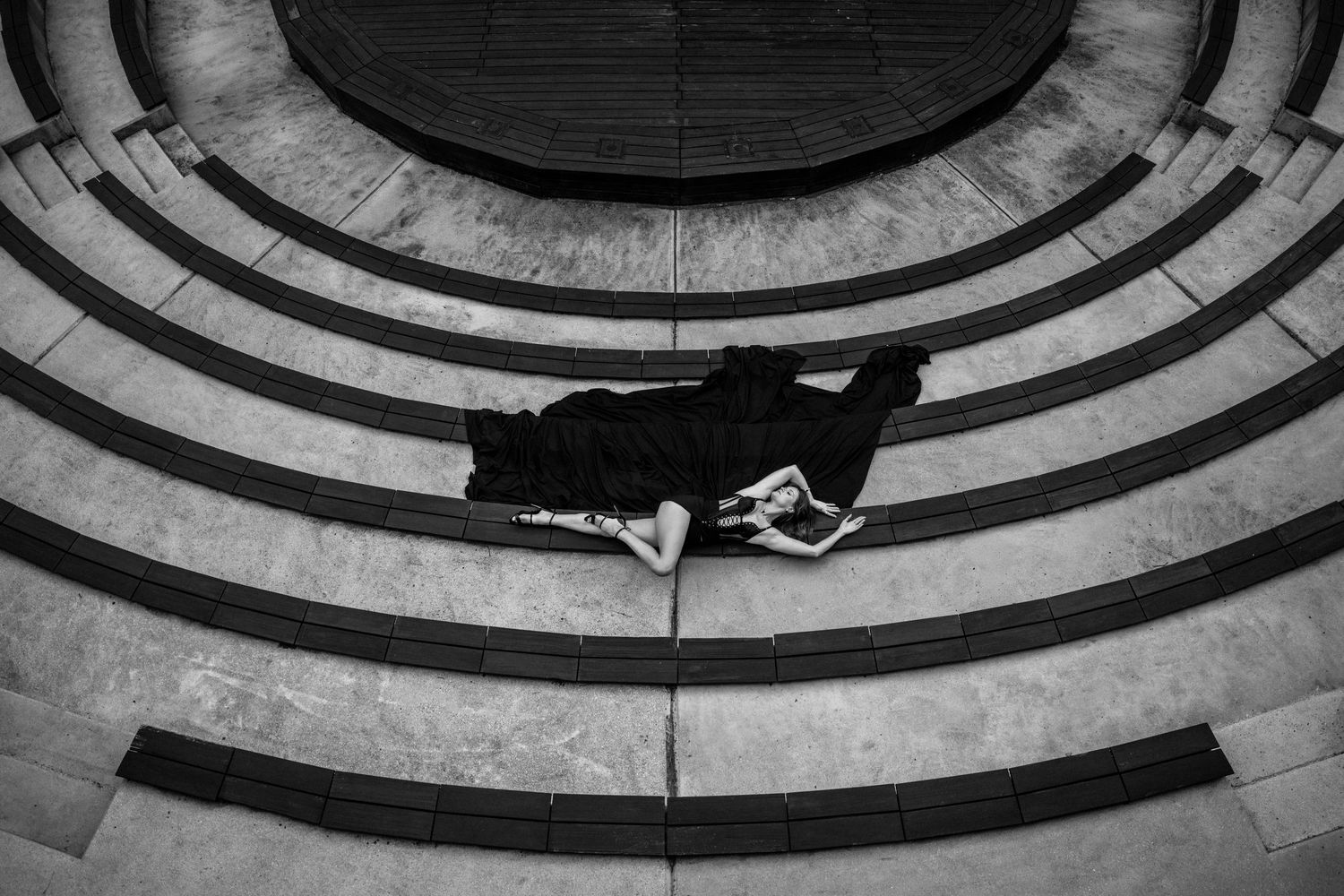Black and white aerial view of a person lying on curved concrete steps in a circular architectural pattern.
