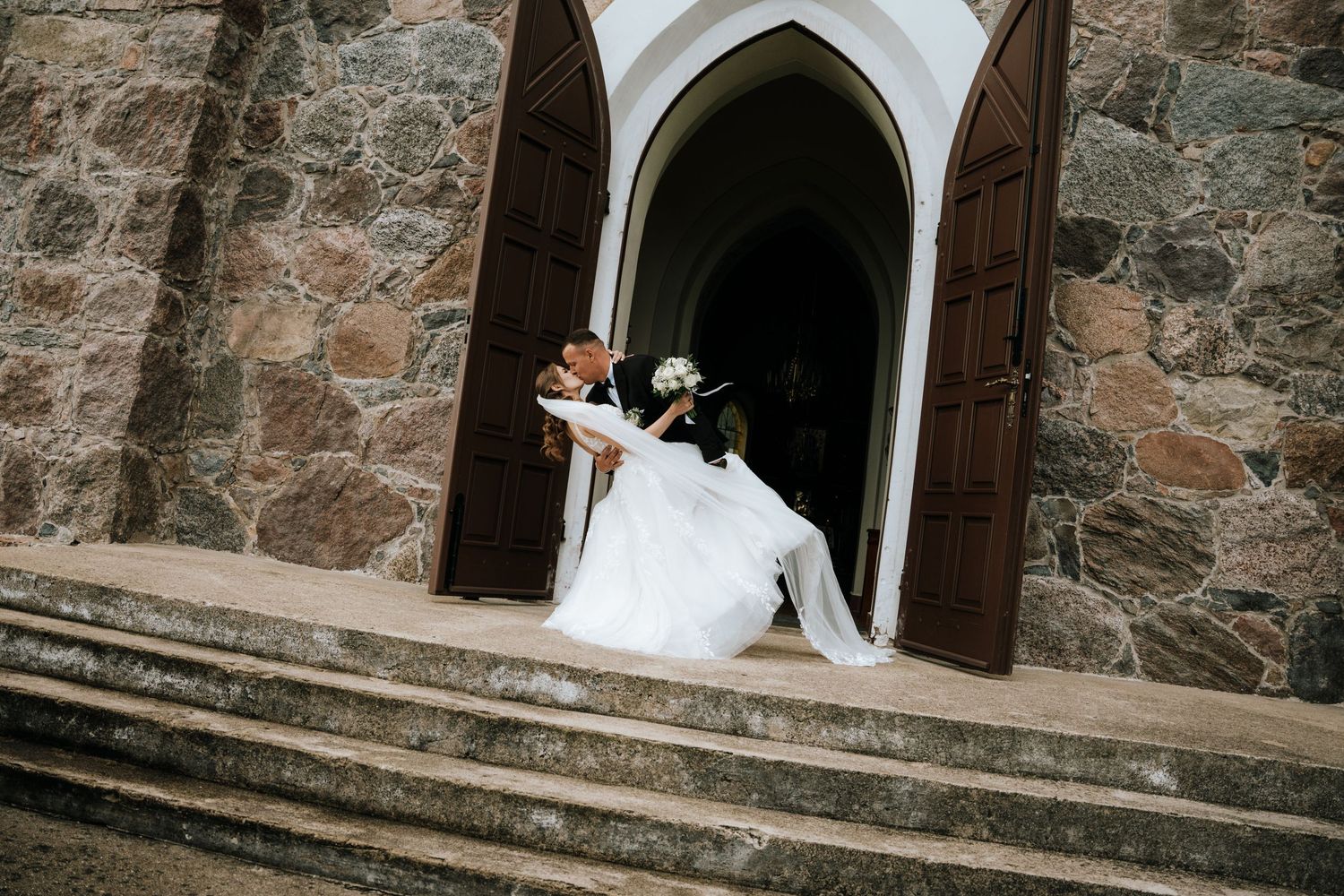 Bride and groom embrace at stone church entrance on wedding day.