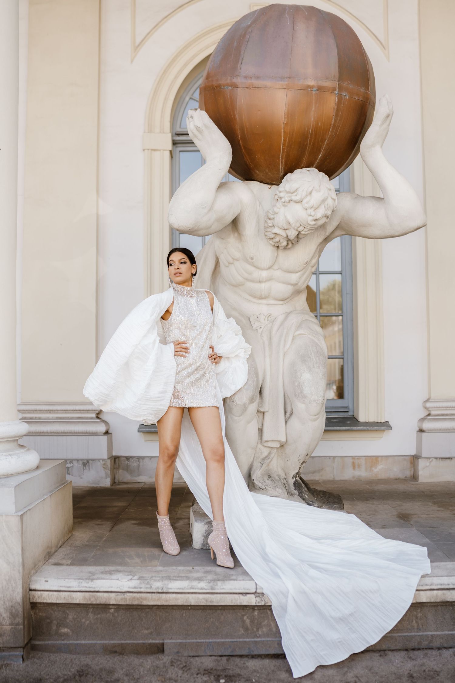 White fashion dress photographed against classical architecture with Atlas statue holding sphere.