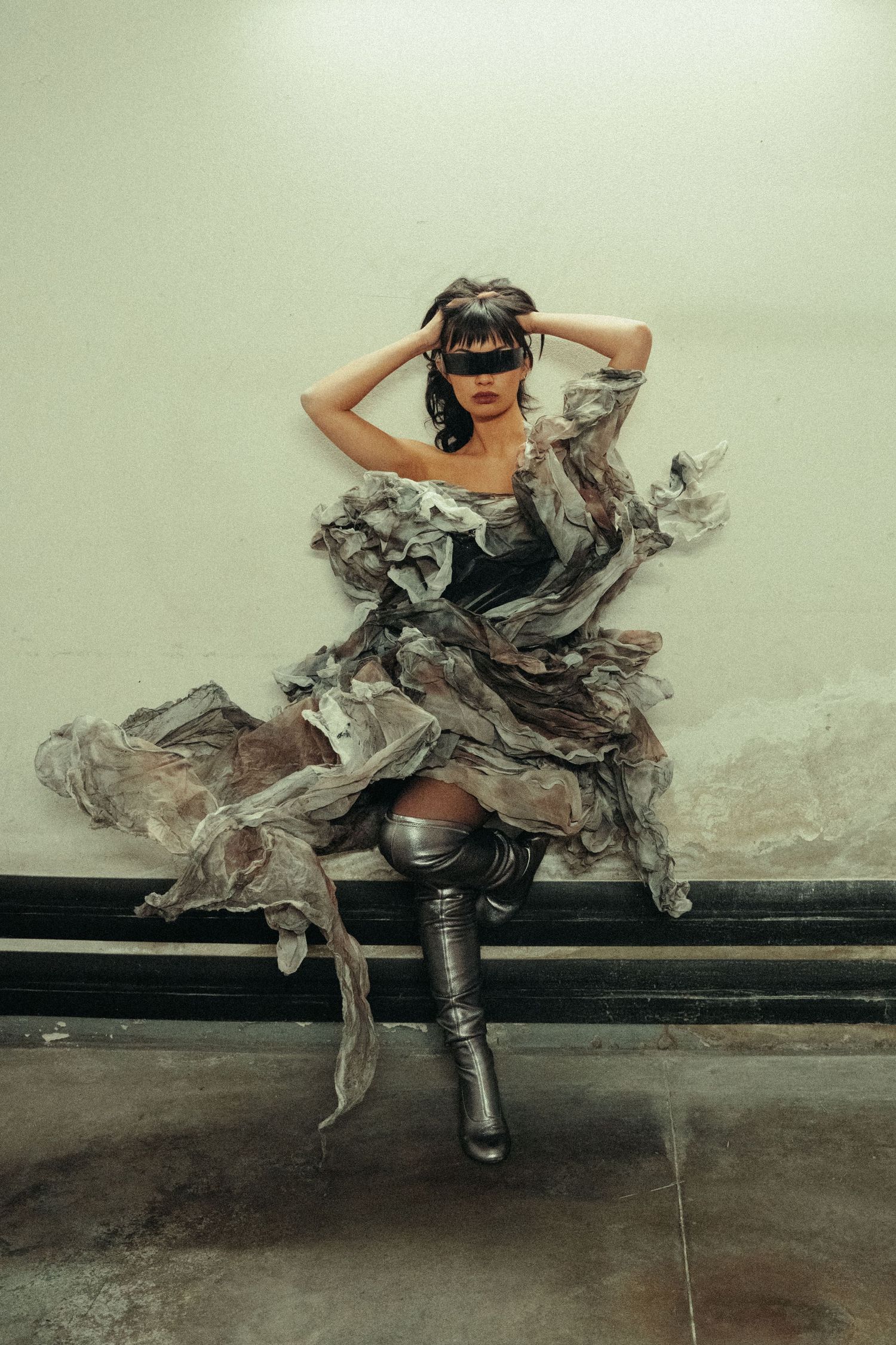 A figure in a dramatic flowing gray dress and tall boots poses artistically against a minimalist wall backdrop.