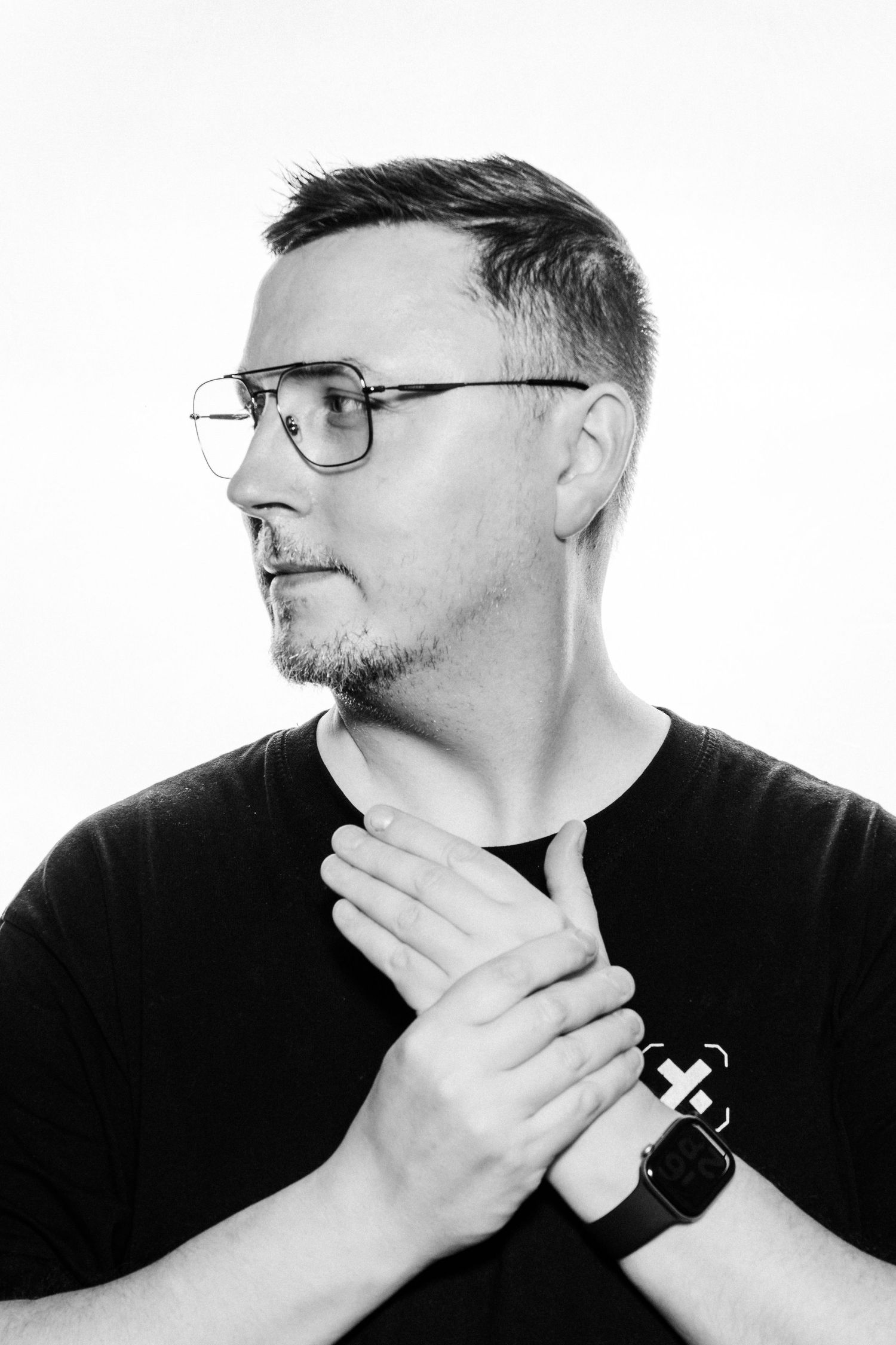 Black and white portrait of a middle-aged man with glasses and stubble, wearing a black t-shirt, hands clasped near chest.