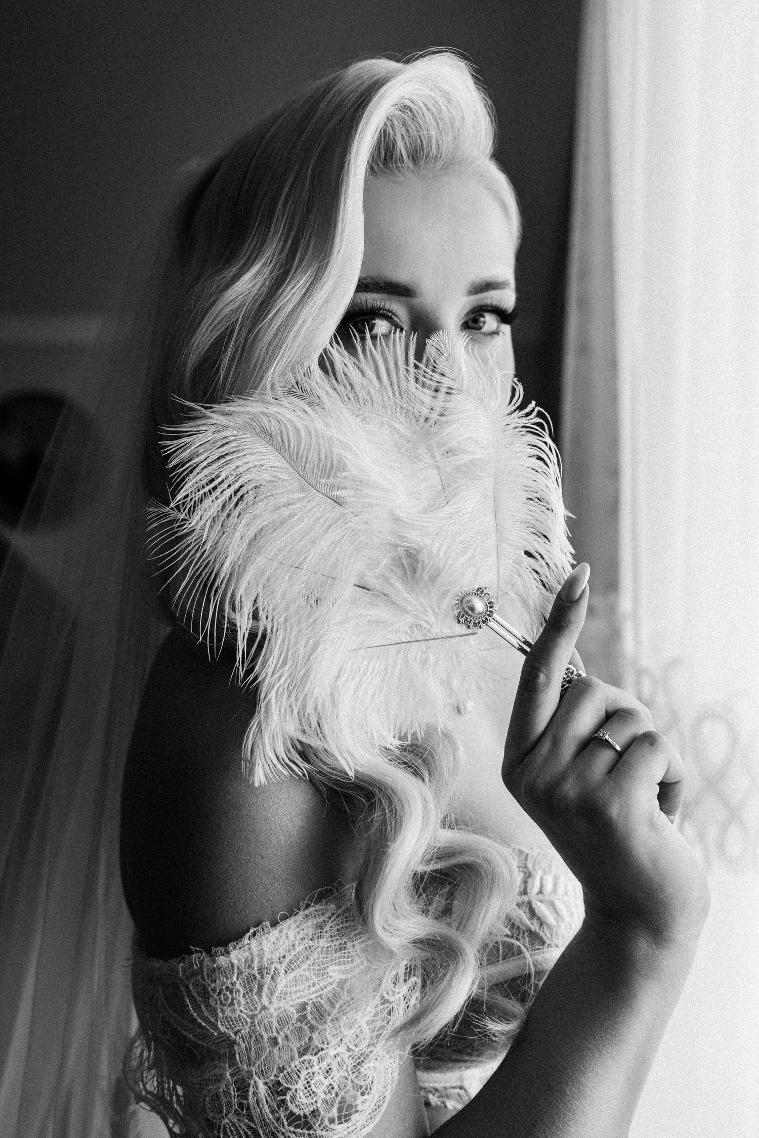 Black and white portrait of a blonde bride holding a feather fan near her face, wearing lace dress and veil.