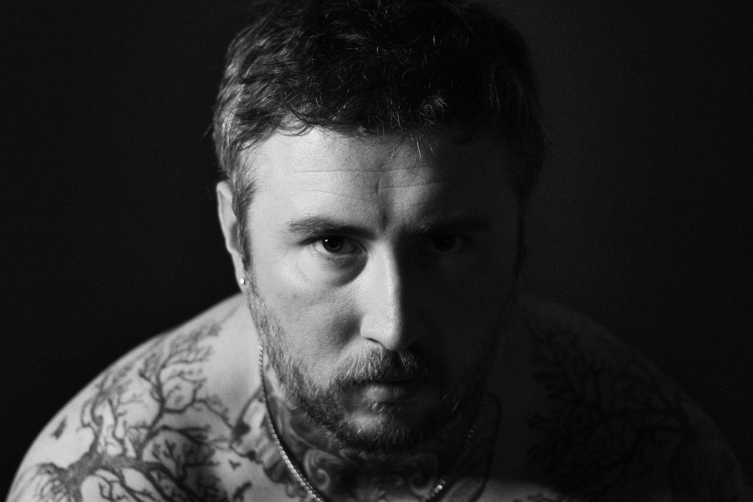High-contrast monochrome portrait highlighting tattoos and expression, captured in a studio setting.
