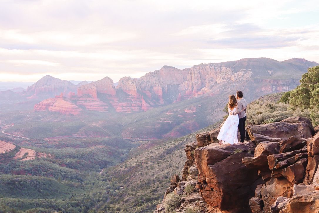 Two figures perch on a dramatic cliff edge overlooking Sedona's vast red rock canyon landscape in Sedona Arizona.