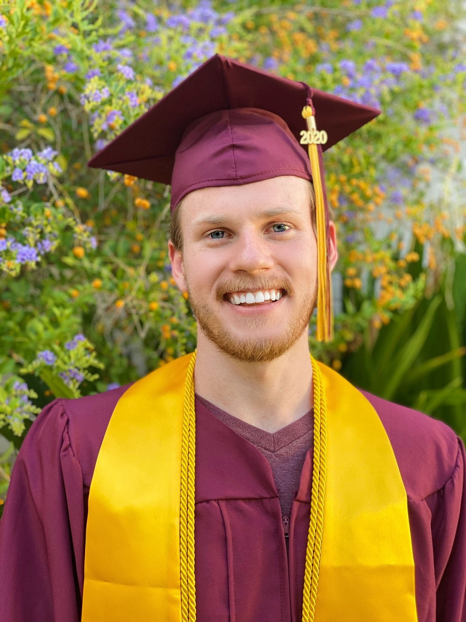 Graduate wearing maroon cap and gown with gold honor cords poses for photos outdoors with flowers and trees in background.
