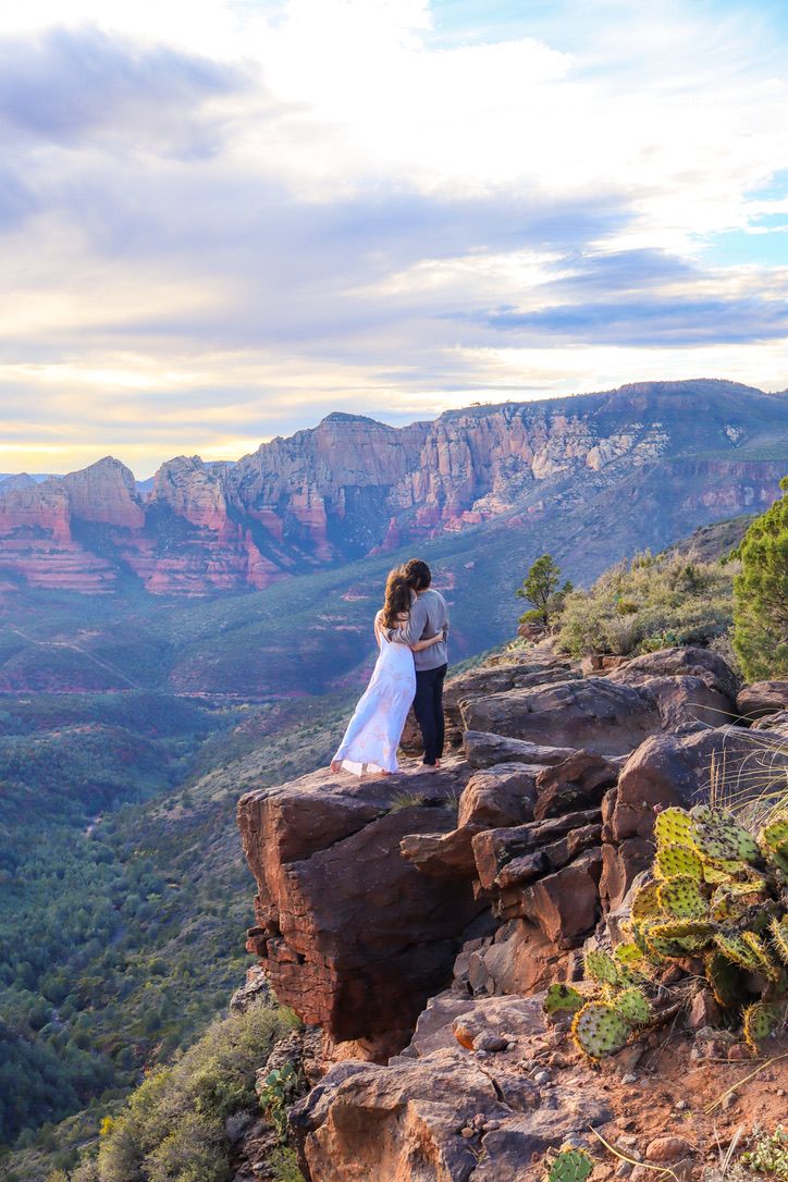 A couple sits on rugged cliffs during sunset with panoramic mountain views in the background in Sedona Arizona.