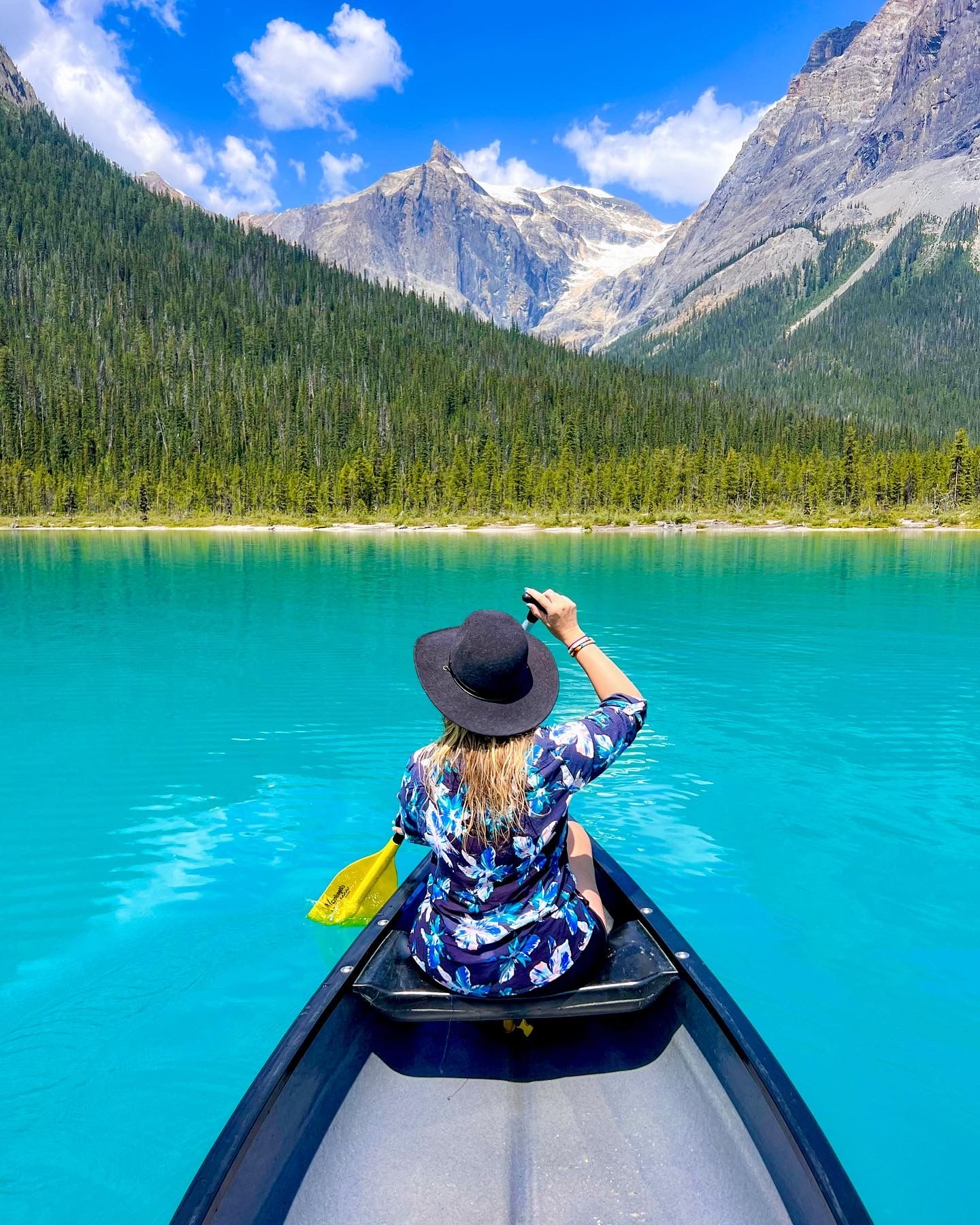 Paddler in a blue shirt and hat canoeing on turquoise alpine lake surrounded by mountain peaks and evergreen forest.