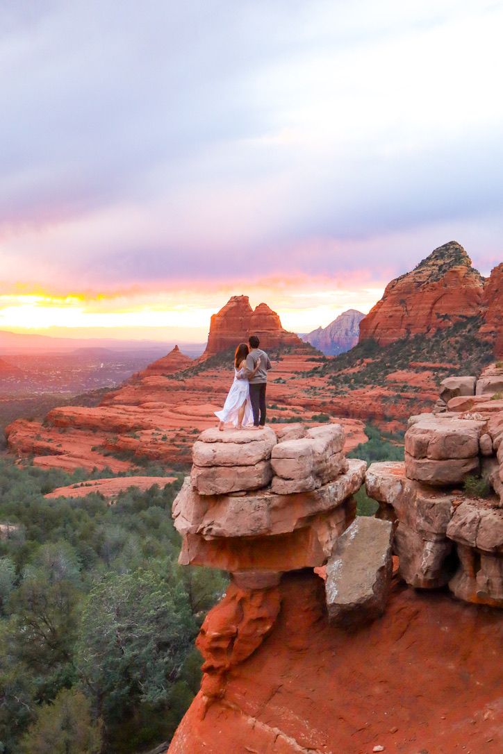 Couple poses on red rock formation with stunning Sedona landscape and colorful sunset sky in Sedona Arizona.