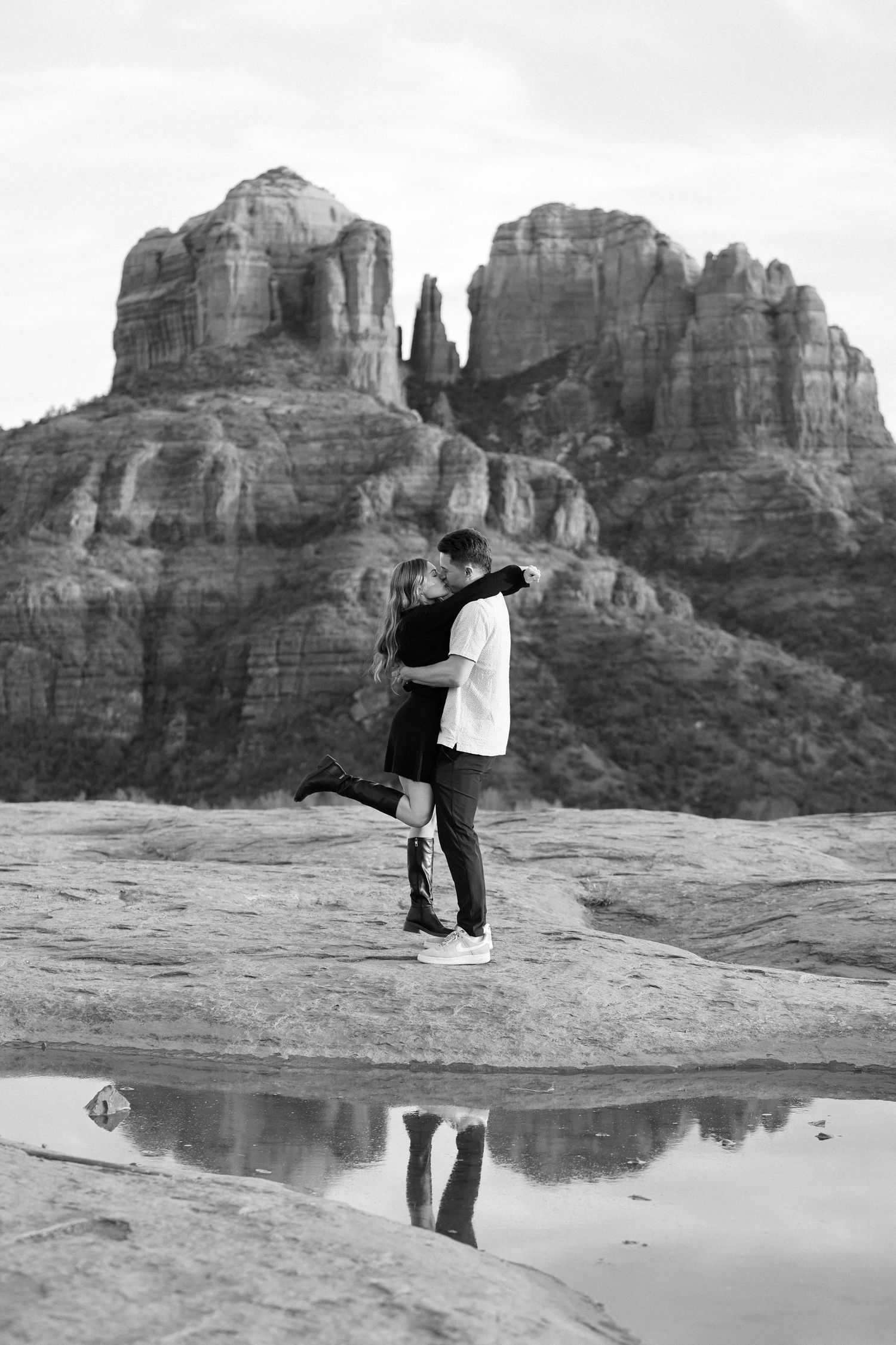 A couple embraces romantically against the dramatic backdrop of red rock formations in Sedona, Arizona with reflective pools.