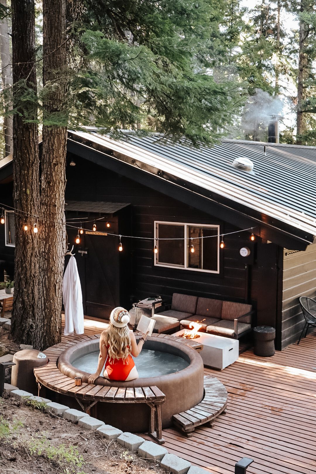 Modern black cabin deck with string lights, wooden benches, and hot tub surrounded by tall pine trees in forest setting.