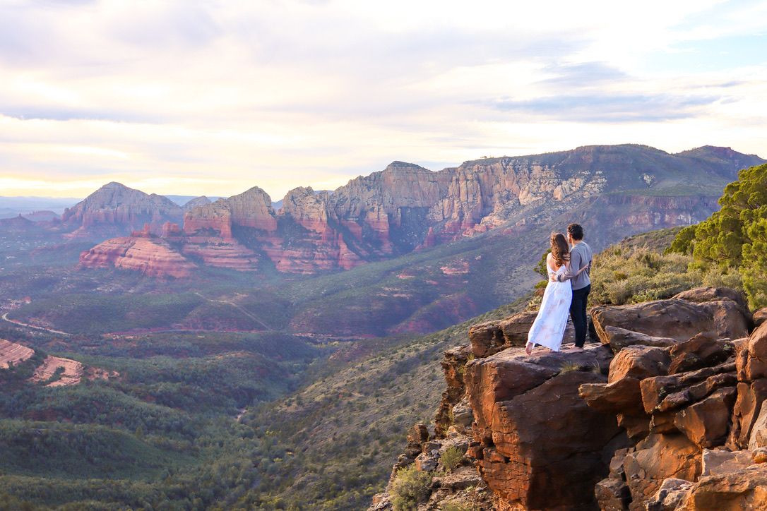 A couple stands on a dramatic red rock cliff overlooking the majestic Sedona valley and mountain range at sunset in Sedona Arizona.