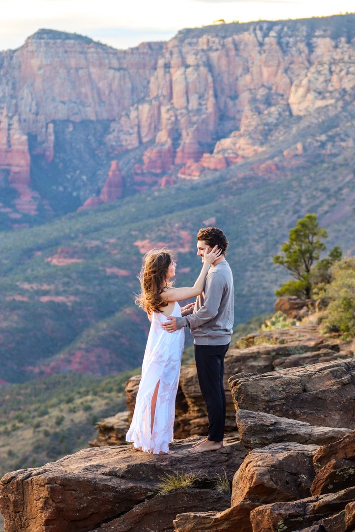 A couple embraces on a rocky outcrop with majestic red cliffs in the background in Sedona Arizona.