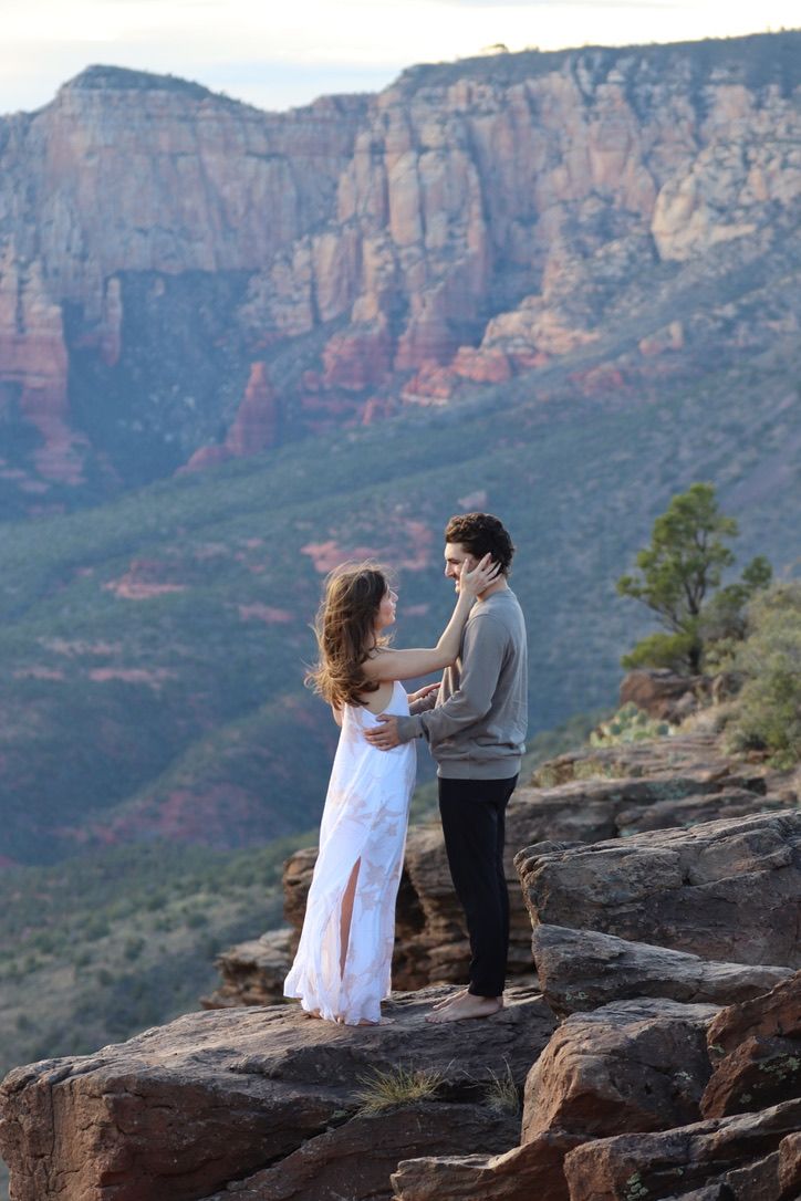 A couple stands on rocky terrain with towering red canyon walls behind them in Sedona Arizona.