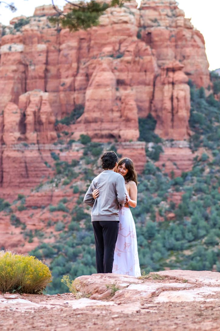 Two people share an intimate moment on a scenic overlook surrounded by towering red cliffs.