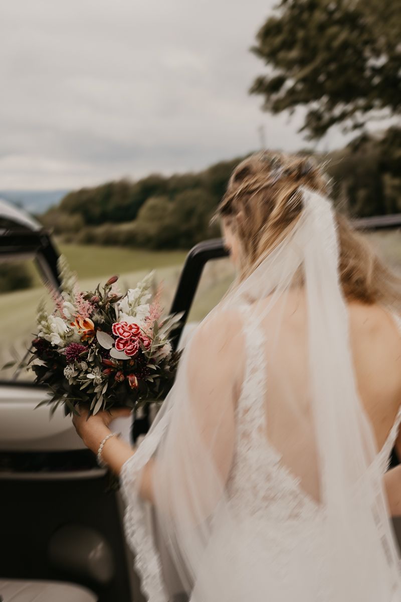 Bride in lace wedding dress and veil holds floral bouquet beside vintage convertible car with scenic green countryside backdrop.