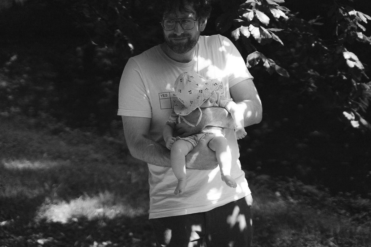 Black and white photo of a bearded man holding a baby outdoors near trees.