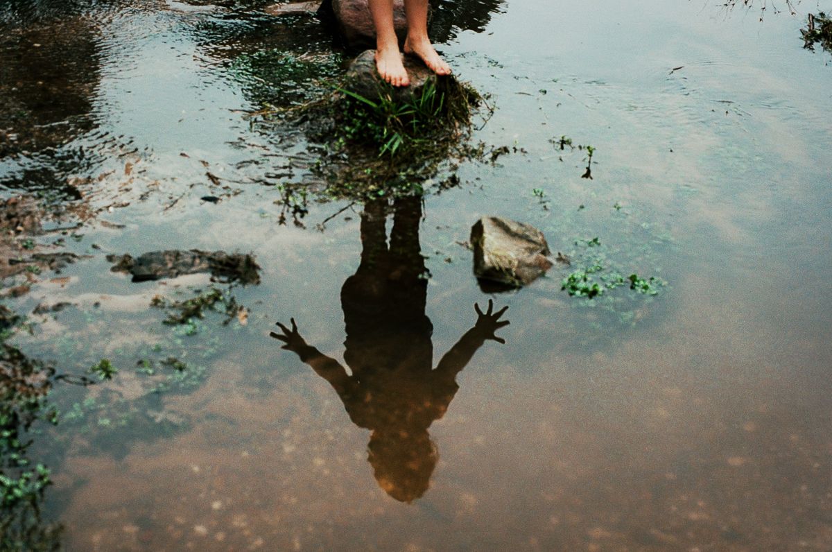 Child standing in shallow water with a shadow reflection visible on the water surface surrounded by rocks and greenery.