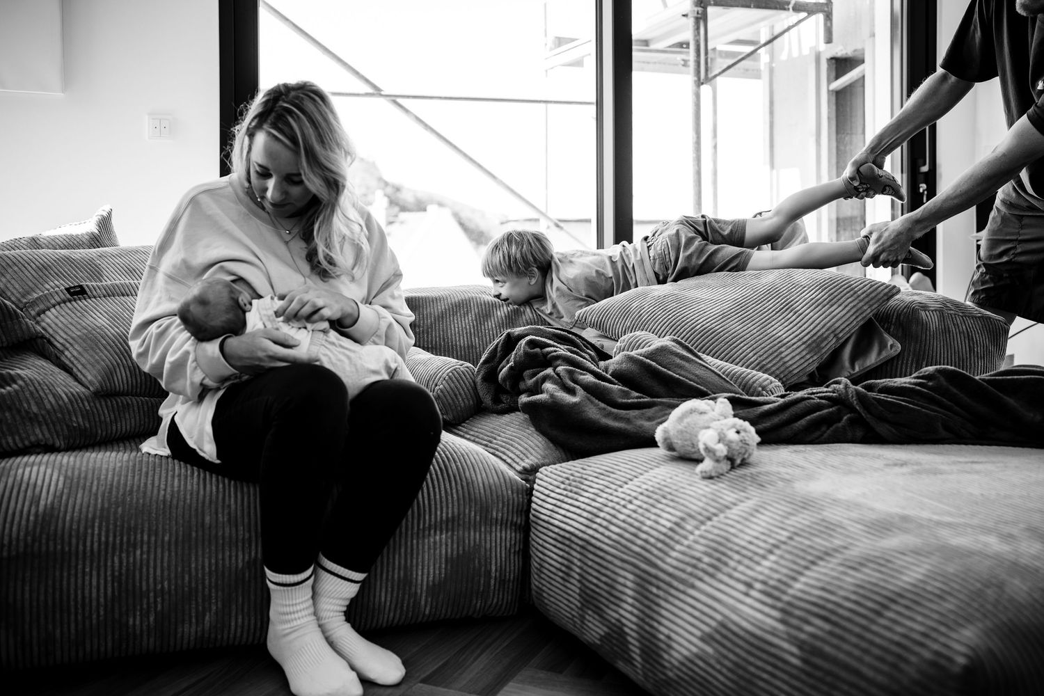 Schwarzweißfoto einer Mutter, die ihr Neugeborenes auf dem Sofa stillt, während ein Kleinkind in der Nähe spielt.