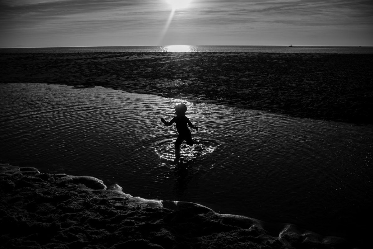 Black and white photo of a child splashing in a shallow beach pool at sunset, silhouetted against the glowing horizon.
