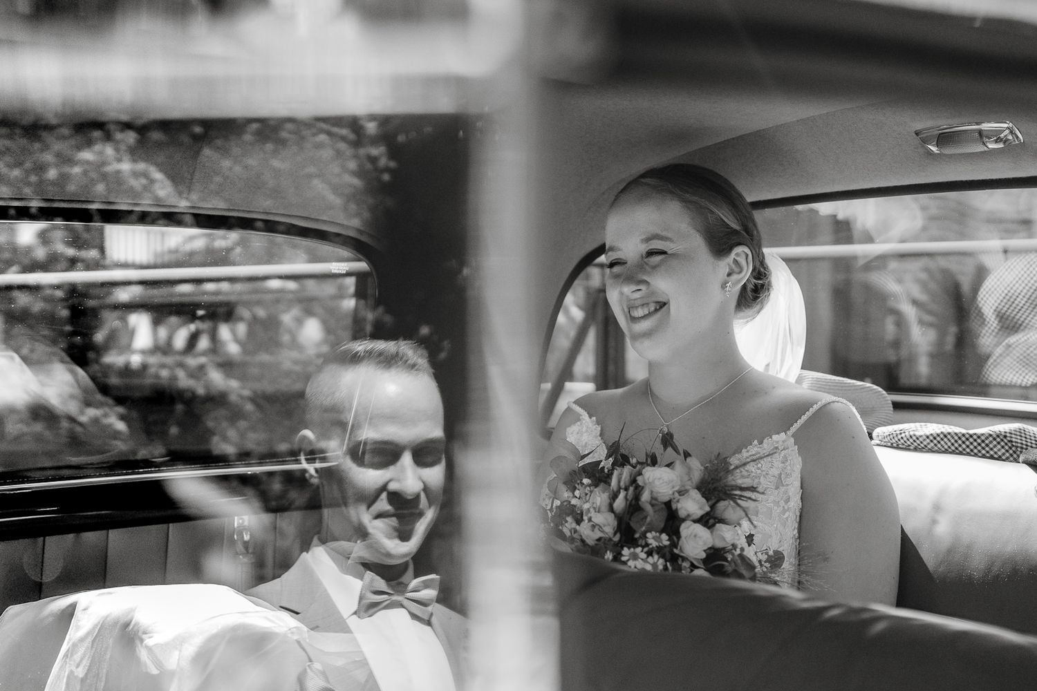 Black and white photo of a smiling bride holding flowers seated in a vintage car with a man in a bow tie beside her.