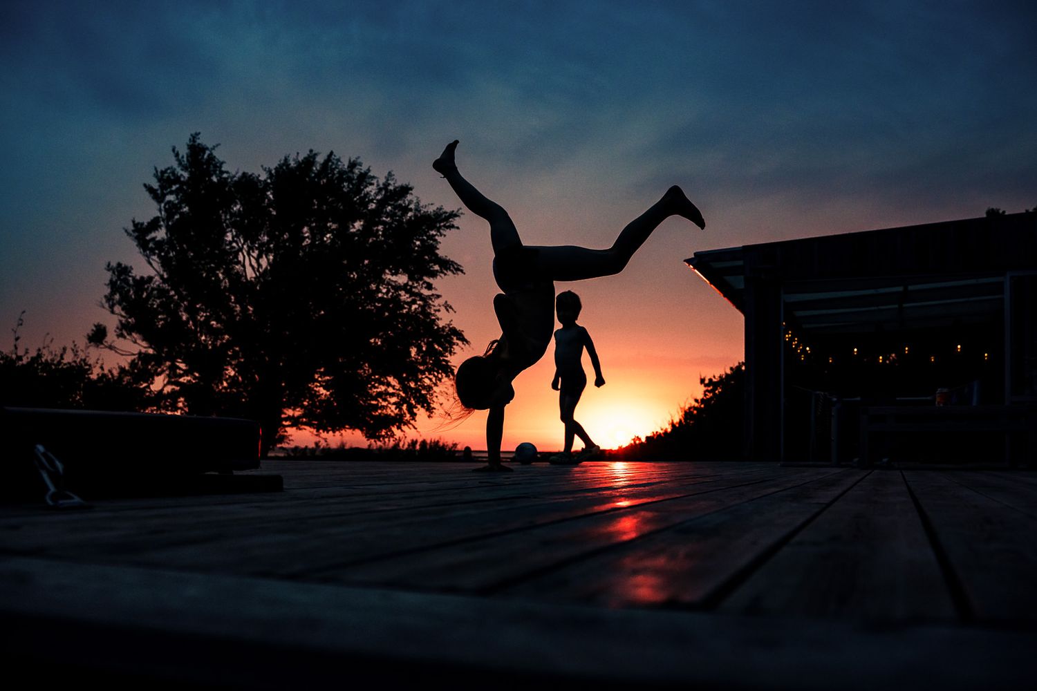 Silhouettes of children playing at sunset, one doing a cartwheel on a wooden deck with glowing horizon behind them.