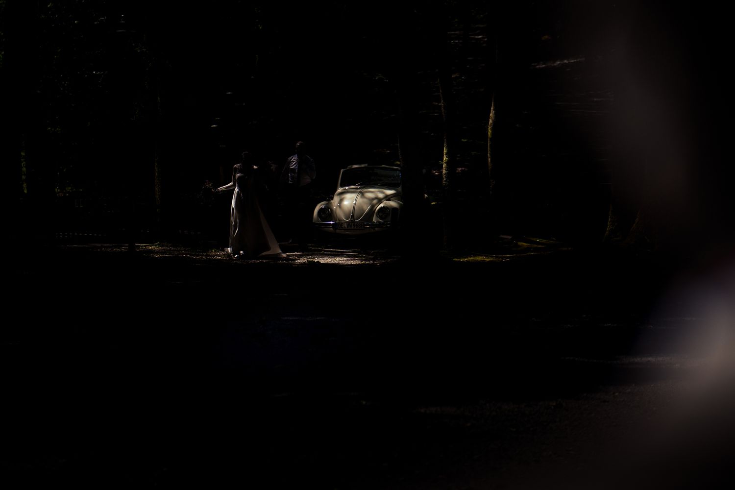 A dimly lit night scene with a vintage car illuminated by a spotlight and a lone figure standing nearby.