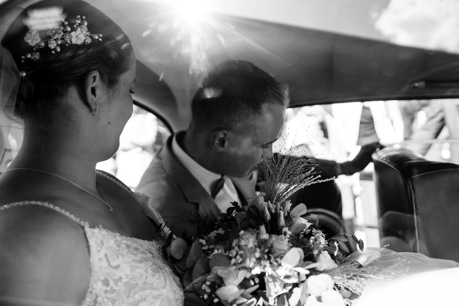 Black and white wedding photo of bride and groom sharing a tender moment in a vintage car with bouquet.