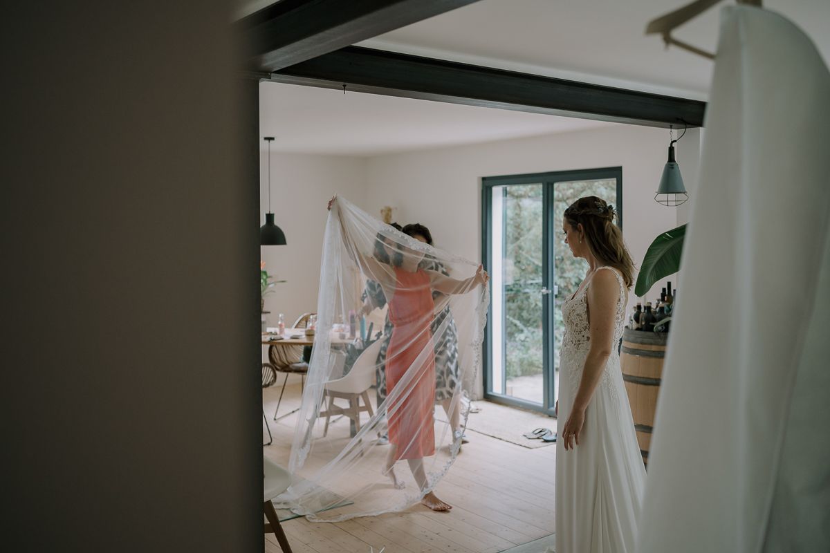 Bridesmaid helping bride with veil in bright rustic room before wedding ceremony.