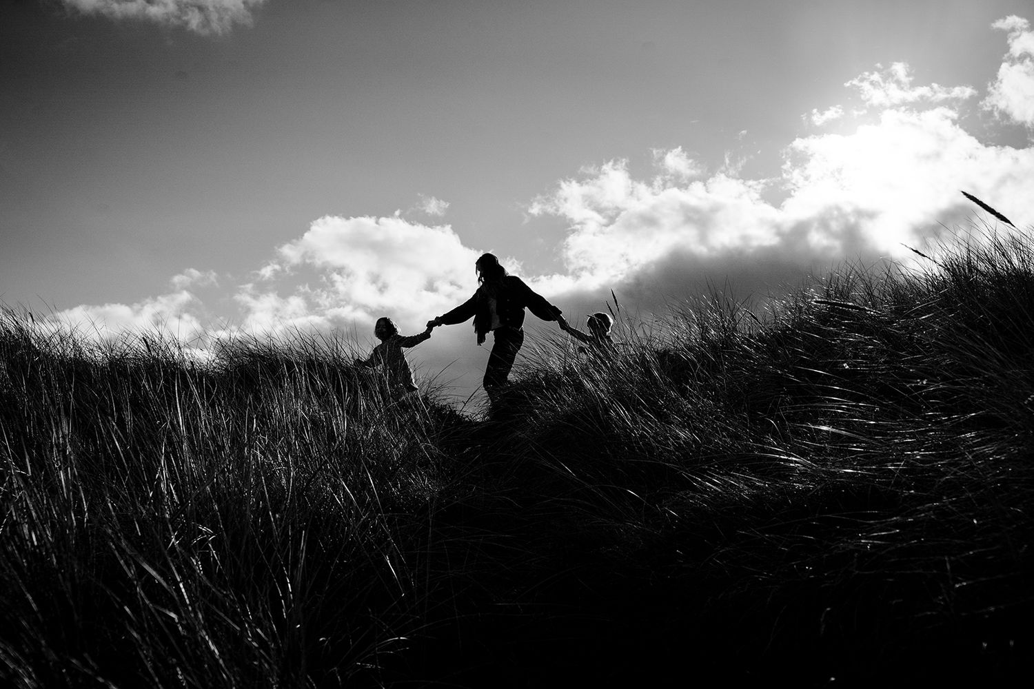 Silhouette of adult and child holding hands climbing grassy hill against dramatic cloudy sky in black and white.